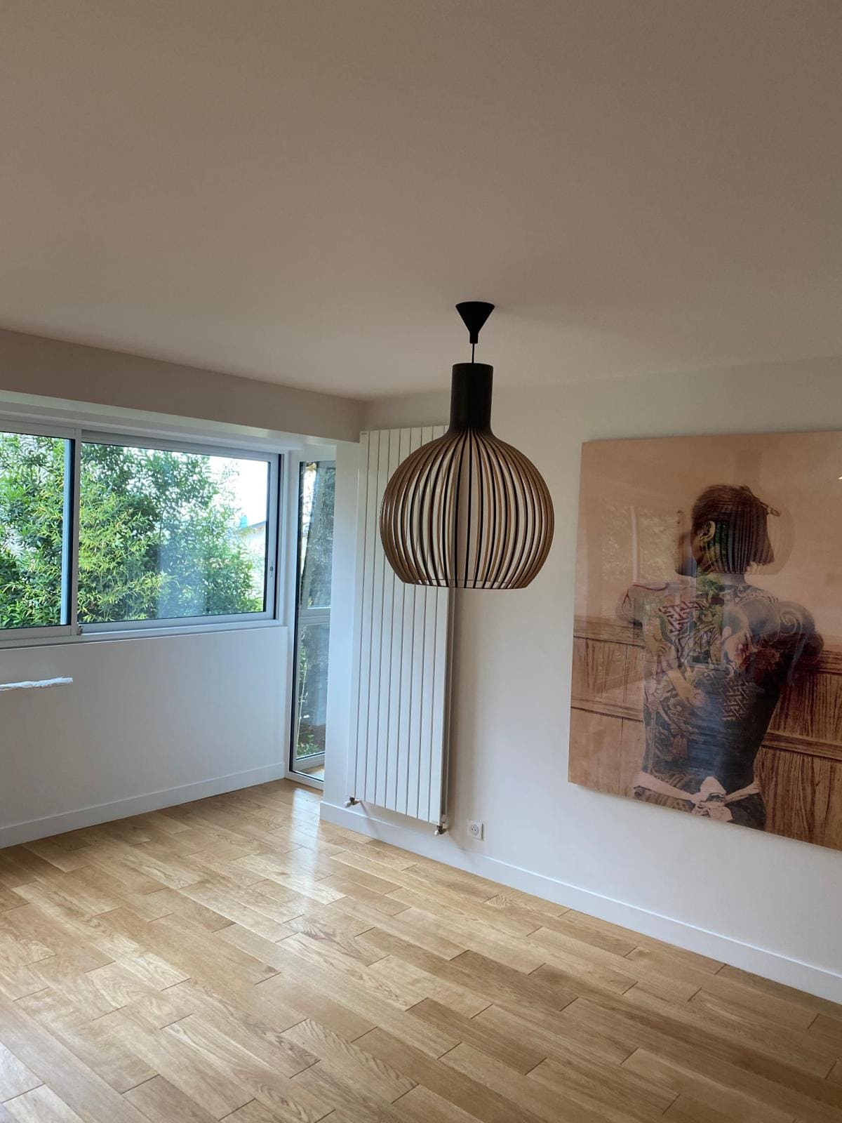 Une pièce non meublé avec un parquet, un radiateur, un tableau, une fenêtre et une lampe suspendue